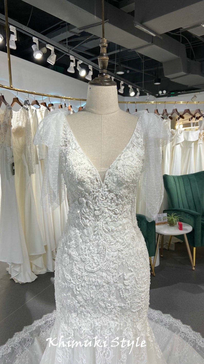 May include: A white wedding dress with a V-neckline and a fitted bodice. The dress has a lace overlay with beaded embellishments and sheer sleeves with bows. The dress is displayed on a mannequin in a store setting.
