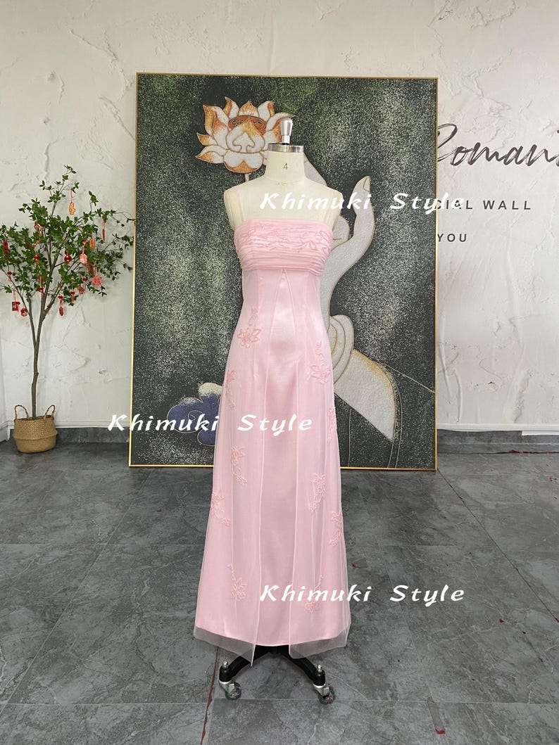 May include: A strapless, floor-length pink dress on a mannequin. The dress features a ruched bodice and embroidered floral details. The skirt is made of a sheer fabric. The words "Khimuki Style" are visible on the dress and in the background.