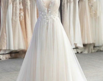 May include: A white tulle wedding dress with a plunging neckline and delicate floral embroidery. The dress has a flowing A-line silhouette and a long train. The dress is displayed on a mannequin in a shop setting.