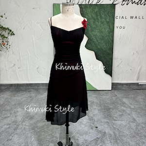 K&ouml;nnte beinhalten: Ein schwarzes, &auml;rmelloses Kleid mit einem figurbetonten Oberteil und einem flie&szlig;enden Rock. Das Kleid hat eine rote Blumenverzierung am Oberteil. Das Kleid wird auf einer Schaufensterpuppe vor einer wei&szlig;en Wand pr&auml;sentiert.