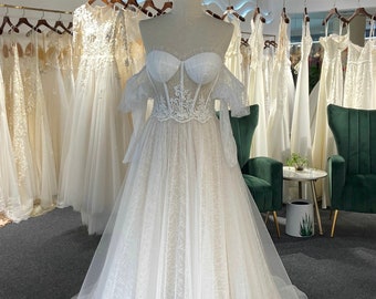 Pode incluir: Vestido de noiva branco em um manequim, com decote em coração, mangas ombro a ombro e corpete de renda. A saia é em estilo A com uma sobreposição estampada. O vestido é exibido em uma loja de noivas com outros vestidos ao fundo. O texto "Khimuki Style" é visível.