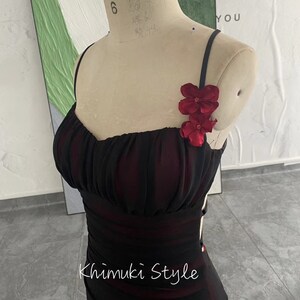 K&ouml;nnte beinhalten: Ein schwarzes Kleid mit einem roten Blumen-Detail am Tr&auml;ger. Das Kleid hat ein figurbetontes Oberteil und einen flie&szlig;enden Rock. Das Kleid befindet sich auf einer Schaufensterpuppe. Der Text "Khimuki Style" ist auf das Kleid gedruckt.