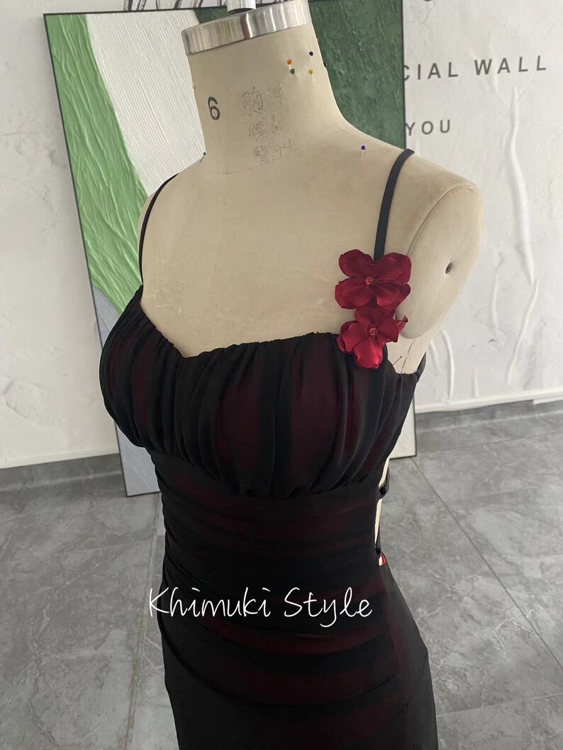 K&ouml;nnte beinhalten: Ein schwarzes Kleid mit einem roten Blumen-Detail am Tr&auml;ger. Das Kleid hat ein figurbetontes Oberteil und einen flie&szlig;enden Rock. Das Kleid befindet sich auf einer Schaufensterpuppe. Der Text "Khimuki Style" ist auf das Kleid gedruckt.