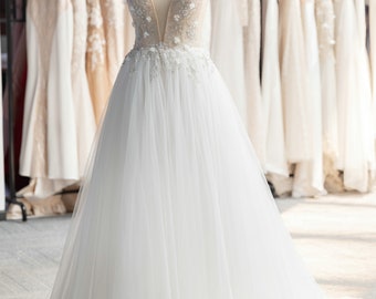 May include: A white tulle wedding dress with a sheer bodice and floral embellishments. The dress has a full skirt and a train. The dress is on a mannequin in a shop.