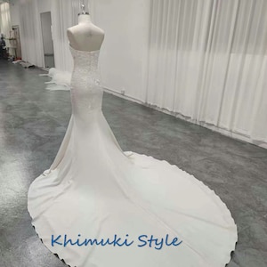 May include: A white strapless wedding dress with a fitted bodice and a flowing skirt. The dress has a lace overlay and a train. The dress is on a mannequin in a white room. The text "Khimuki Style" is printed on the skirt.