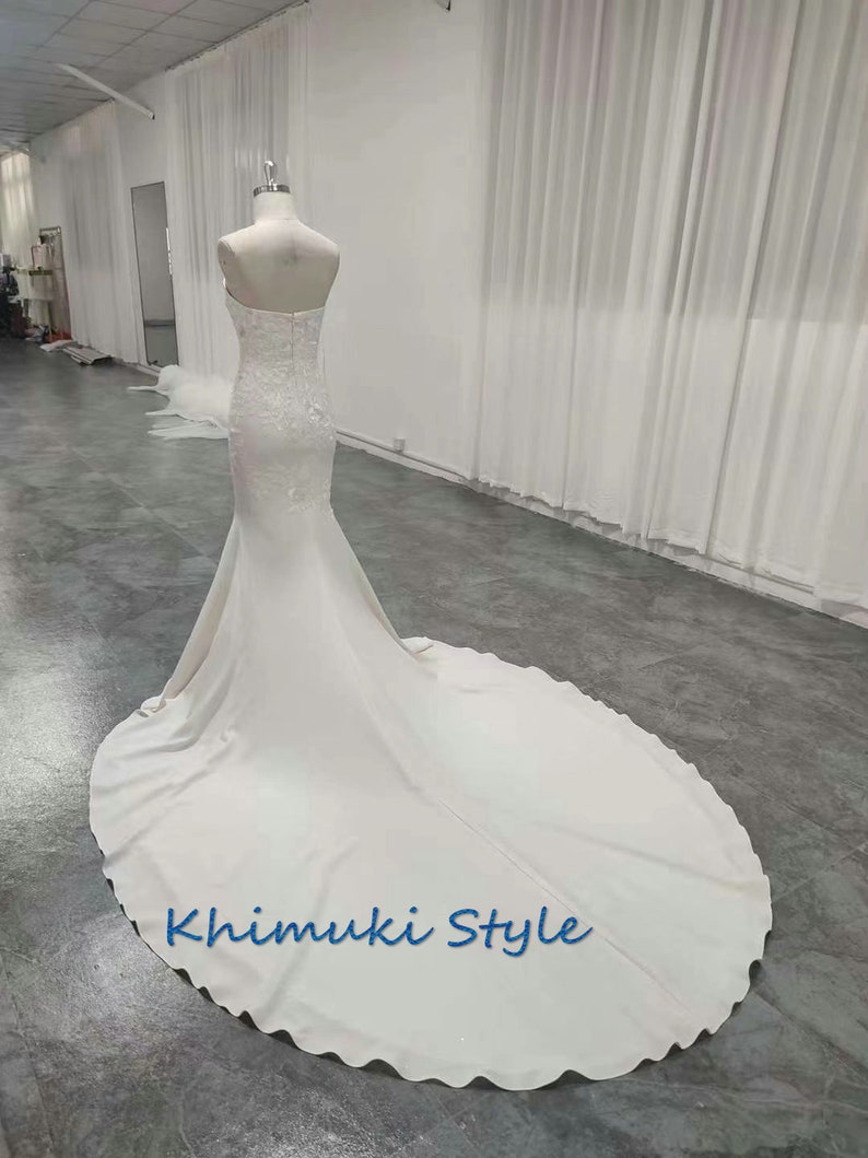 May include: A white strapless wedding dress with a fitted bodice and a flowing skirt. The dress has a lace overlay and a train. The dress is on a mannequin in a white room. The text "Khimuki Style" is printed on the skirt.