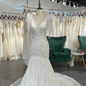 May include: A white lace wedding dress with a V-neckline and a fitted bodice. The dress has a long train and a lace overlay. The dress is displayed on a mannequin in a bridal shop.