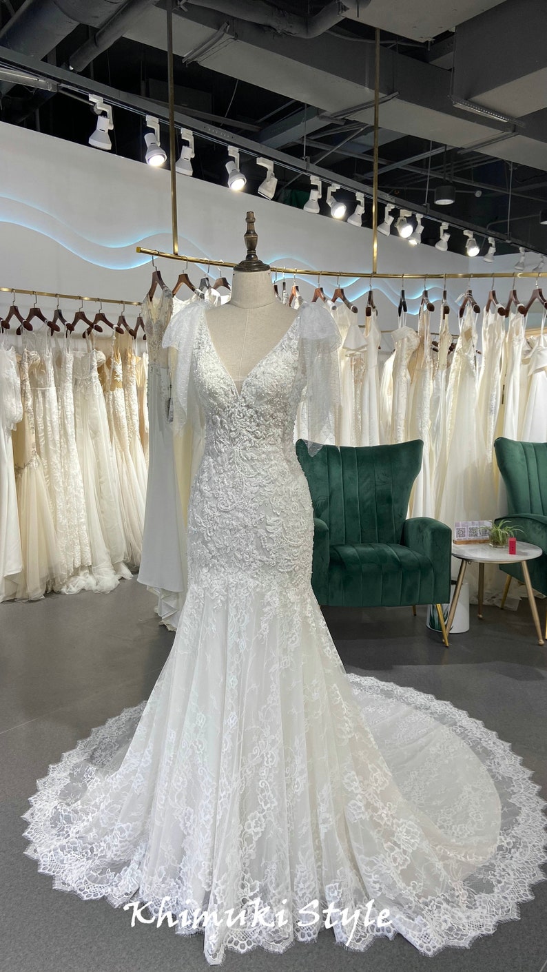 May include: A white lace wedding dress with a V-neckline and a fitted bodice. The dress has a long train and a lace overlay. The dress is displayed on a mannequin in a bridal shop.