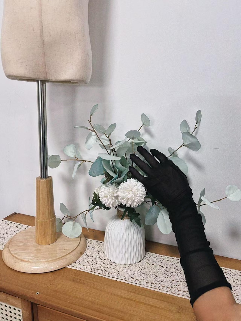 May include: A display featuring a beige dress form, a white vase with white flowers and greenery, and a pair of long black sheer gloves. The vase sits on a wooden table with a lace runner.