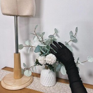 K&ouml;nnte beinhalten: Ein Paar schwarze, durchsichtige Handschuhe mit einer wei&szlig;en Vase mit Blumen im Hintergrund.