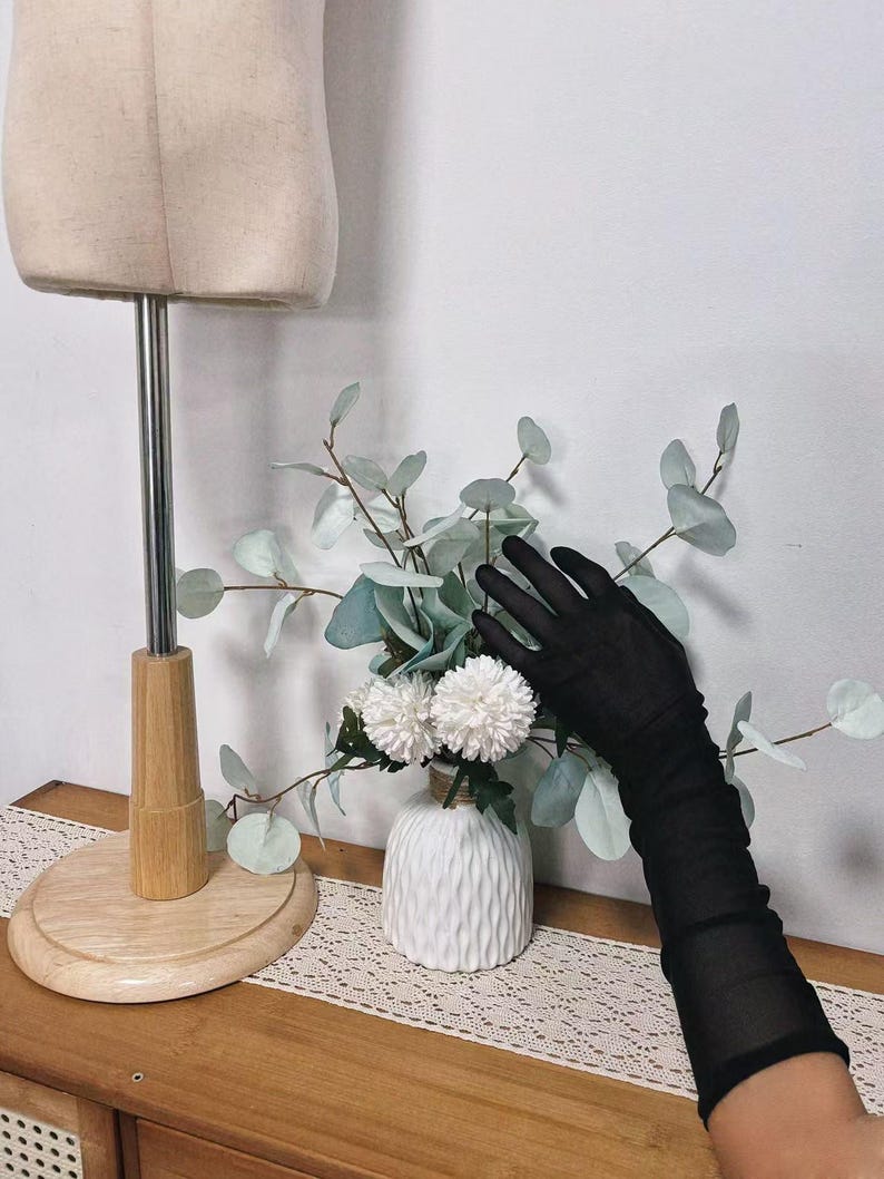 K&ouml;nnte beinhalten: Ein Paar schwarze, durchsichtige Handschuhe mit einer wei&szlig;en Vase mit Blumen im Hintergrund.