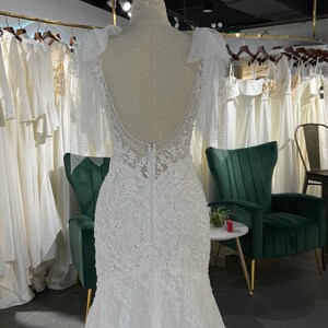 May include: A white lace wedding dress with a low back and a fitted bodice. The dress has a long train and is decorated with delicate beading. The dress is displayed on a mannequin in a store.