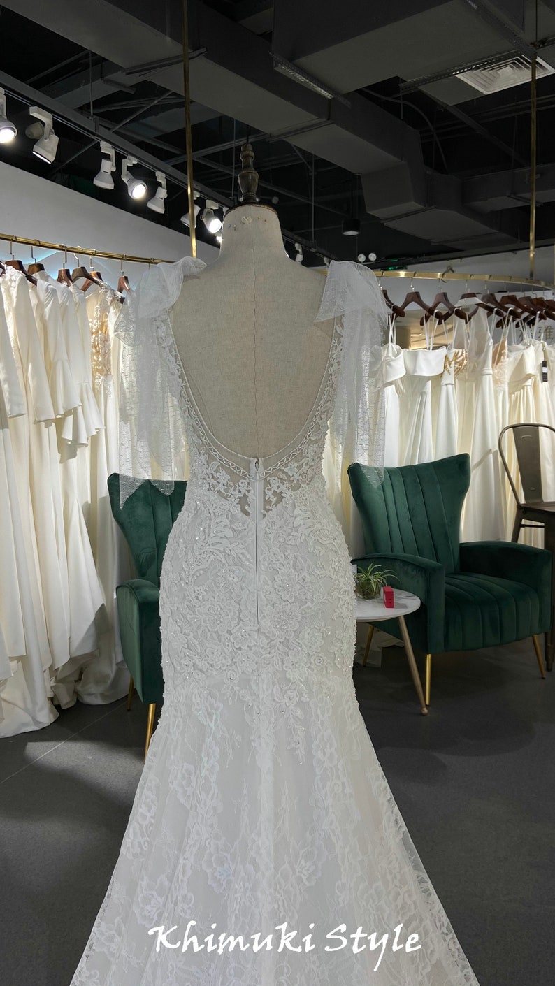 May include: A white lace wedding dress with a low back and a fitted bodice. The dress has a long train and is decorated with delicate beading. The dress is displayed on a mannequin in a store.