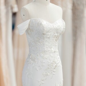 May include: A white off-the-shoulder wedding dress with a fitted bodice and a flowing skirt. The dress is adorned with delicate lace and beading.