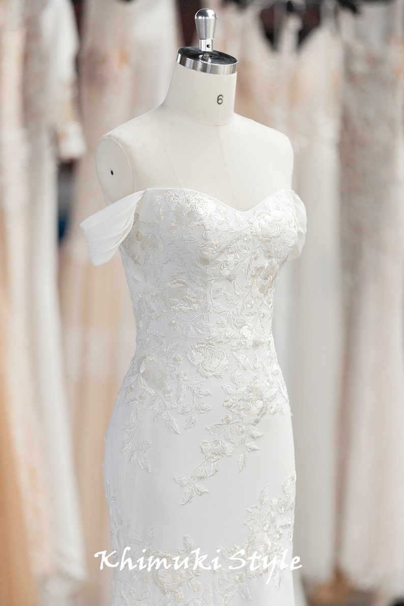 May include: A white off-the-shoulder wedding dress with a fitted bodice and a flowing skirt. The dress is adorned with delicate lace and beading.