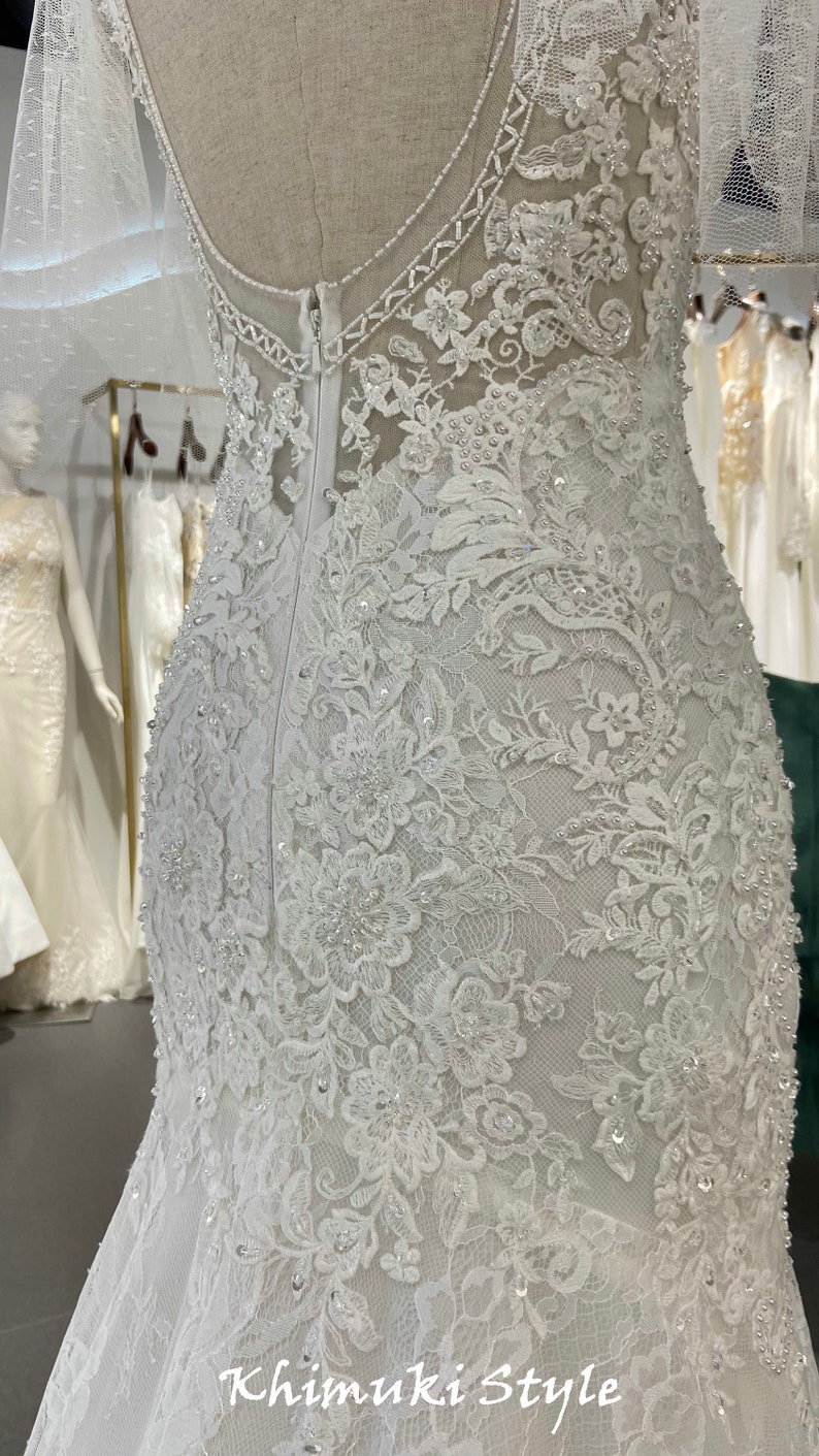 May include: A white lace wedding dress with a fitted bodice and a flowing skirt. The dress is decorated with delicate beading and floral patterns. The back of the dress features a zipper closure and a low back.