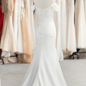 May include: A white off-the-shoulder mermaid wedding dress with delicate lace detailing and a flowing train. The dress is displayed on a mannequin in a shop setting.