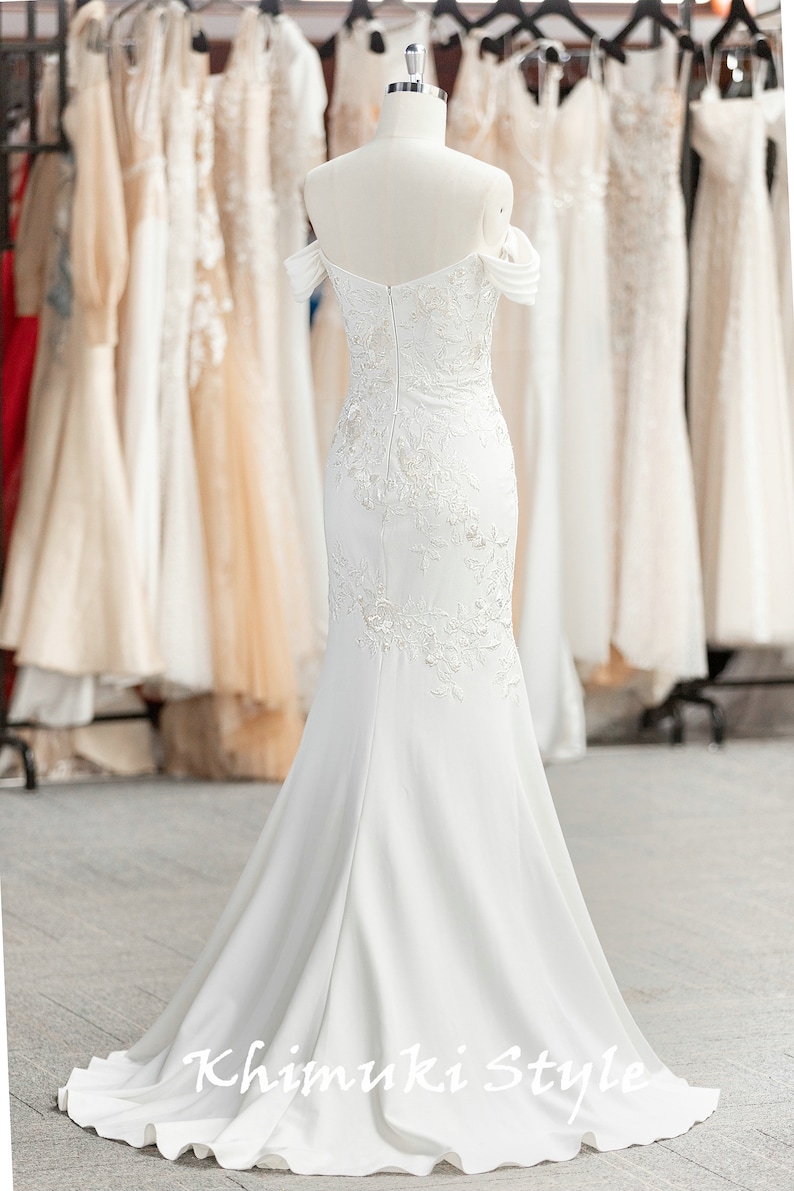 May include: A white off-the-shoulder mermaid wedding dress with delicate lace detailing and a flowing train. The dress is displayed on a mannequin in a shop setting.