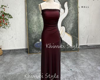 Pode incluir: Um vestido maxi cor vinho com um corpete justo e uma saia fluida. O vestido tem um decote quadrado e alças finas. O vestido está em um manequim e é exibido contra uma parede branca com uma pintura no fundo.
