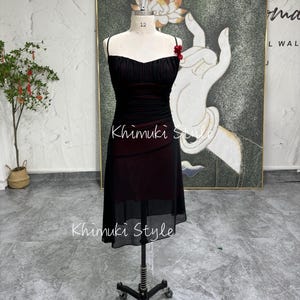 K&ouml;nnte beinhalten: Ein schwarzes und rotes &auml;rmelloses Kleid mit einem herzf&ouml;rmigen Ausschnitt und einem gerafften Rock. Das Kleid befindet sich auf einer Schaufensterpuppe und hat eine rote Blumenverzierung am Tr&auml;ger.