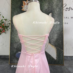 May include: A light pink dress on a mannequin with a lace-up back. The dress has spaghetti straps and a flowing skirt. The text "Khimuki Style" is visible on the dress. The dress is displayed in front of a painting with a floral design.