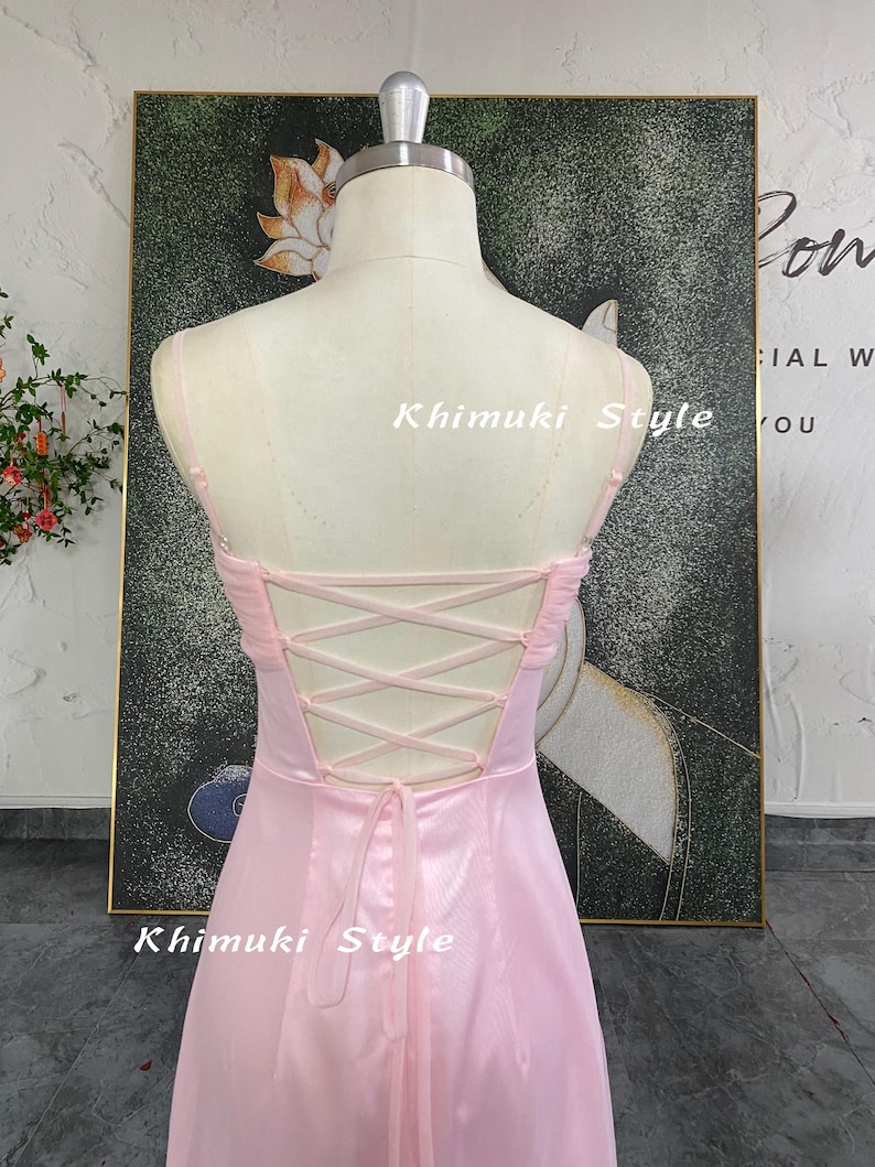 May include: A light pink dress on a mannequin with a lace-up back. The dress has spaghetti straps and a flowing skirt. The text "Khimuki Style" is visible on the dress. The dress is displayed in front of a painting with a floral design.
