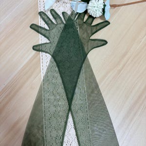 May include: A pair of long, green, sheer gloves with delicate lace detailing. The gloves are laid out on a white lace surface.