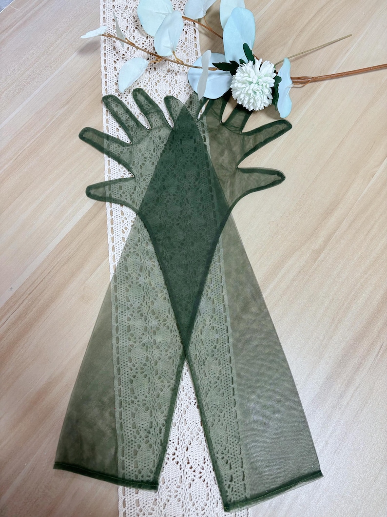 May include: A pair of long, green, sheer gloves with delicate lace detailing. The gloves are laid out on a white lace surface.