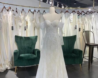 May include: A white lace wedding dress with a sweetheart neckline and off-the-shoulder sleeves. The dress has a fitted bodice and a flowing skirt. The dress is displayed on a mannequin in a bridal shop.