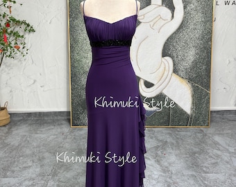 May include: A purple floor-length dress with a black beaded waistband and a ruffled hem. The dress has a sweetheart neckline and spaghetti straps. The dress is on a mannequin in front of a painting.