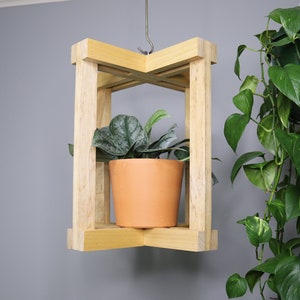 May include: A wooden hanging plant shelf with a single potted plant. The shelf is made of light brown wood and has a simple, modern design. The plant is a small, leafy plant with green leaves.