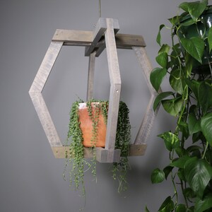 Puede incluir: Un macetero colgante de madera gris con un diseño geométrico. Contiene una maceta de terracota con una planta colgante. El marco de madera está construido con piezas angulares, creando una forma hexagonal. El follaje verde de la planta contrasta con la madera gris y la terracota.