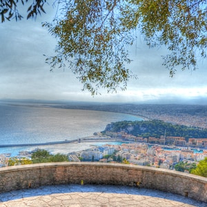 Puede incluir: Una vista panorámica desde un mirador de piedra con vistas a una ciudad y el océano. La ciudad está construida en una península con un puerto y un rompeolas. El océano es de color azul claro y el cielo es de un azul brumoso.
