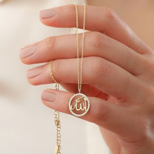 May include: A gold-colored necklace featuring a pendant with the Arabic word "Allah" inscribed within a circle of small, clear stones. The necklace is held up, showcasing its delicate chain and pendant design.