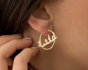 May include: Gold-colored hoop earrings with the Arabic name "عائشة" (Aisha) elegantly integrated into the design. The earrings are circular, with the name's letters forming a stylish accent. The earrings are worn on an ear.