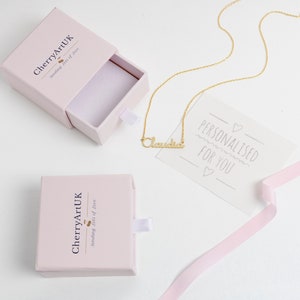 May include: A gold-colored necklace with the name "Claudia" in cursive script, displayed on a white card that reads "Personalised for You." The necklace is next to a pink gift box with the logo "CherryArtUK" and a pink ribbon.