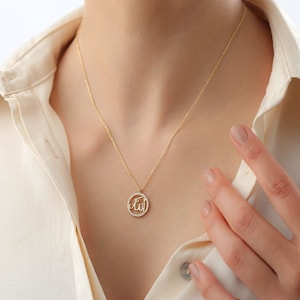 May include: A gold-colored necklace with a pendant featuring the Arabic word for God, "Allah," in a circular design. The pendant is encrusted with small, clear stones. The necklace is worn against a cream-colored shirt.