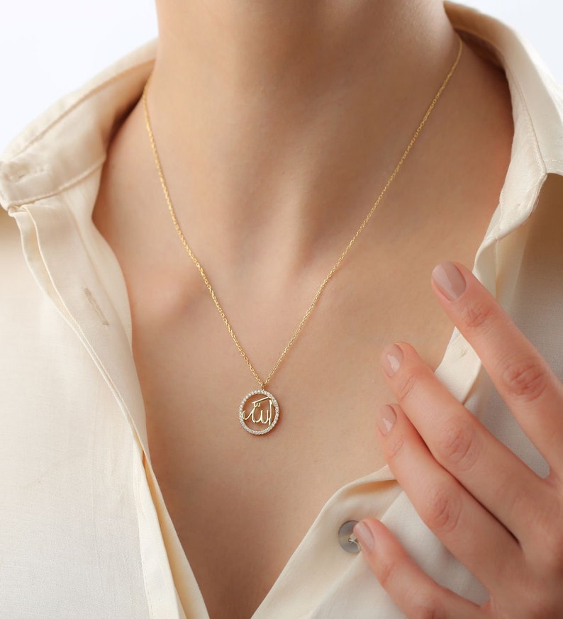 May include: A gold-colored necklace with a pendant featuring the Arabic word for God, "Allah," in a circular design. The pendant is encrusted with small, clear stones. The necklace is worn against a cream-colored shirt.