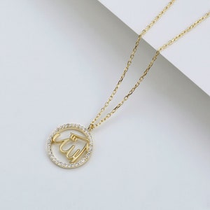 May include: A gold-colored necklace featuring a pendant with the Arabic script for "Allah" encircled by small, clear gemstones. The pendant hangs from a delicate gold chain, set against a white background.