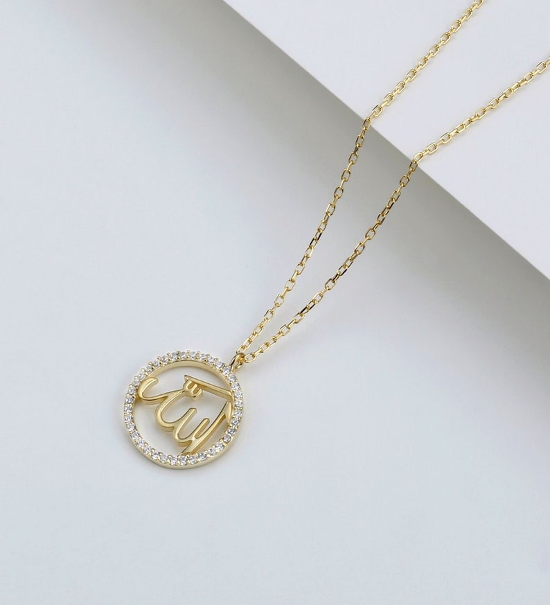 May include: A gold-colored necklace featuring a pendant with the Arabic script for "Allah" encircled by small, clear gemstones. The pendant hangs from a delicate gold chain, set against a white background.