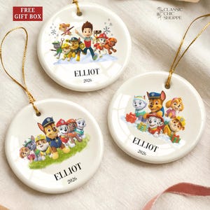 May include: Three white ceramic Christmas ornaments with gold string hangers. Each ornament features a colorful illustration of the Paw Patrol characters and the name "ELLIOT" with the year "2026". The ornaments are round and have a glossy finish.