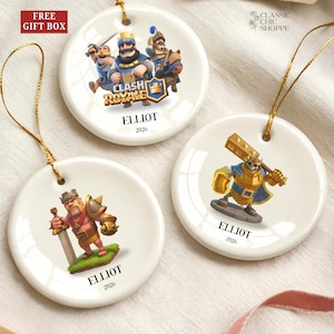 May include: Three white ceramic Christmas ornaments with gold string hangers. Each ornament features a Clash Royale character design, the name "ELLIOT", and the year "2026". The ornaments are round and have a glossy finish.