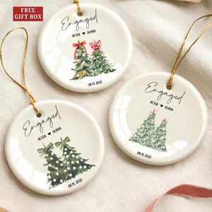 May include: Three white ceramic engagement ornaments with gold string. Each ornament features a hand-painted Christmas tree design and the words "Engaged," the couple's names, and the date 08.15.2025. The top left corner reads "FREE GIFT BOX."