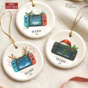 May include: Three white ceramic Christmas ornaments with gold string. Each ornament features a video game controller design with holiday decorations, the name "ELLIOT", and the year "2026". One ornament has a Santa hat.
