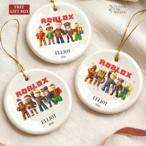 May include: Three white ceramic Christmas ornaments with gold string. Each ornament features the word "ROBLOX" in red, along with colorful character illustrations and the name "ELLIOT" and the year "2026".
