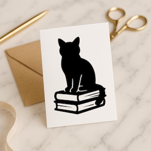 May include: A white card featuring a black silhouette of a cat sitting atop two stacked books. A gold pen, scissors, and a brown envelope are in the background, suggesting a stationery or greeting card theme.