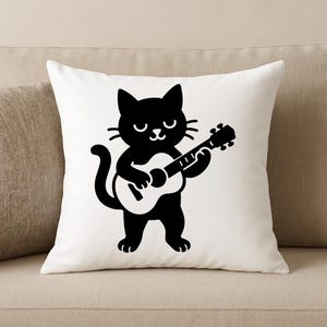May include: White throw pillow featuring a black cartoon cat playing a guitar. The cat has a smiling face and is holding the guitar with both paws. The pillow is square and sits on a beige sofa.