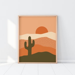 May include: A framed art print featuring a desert landscape with a cactus silhouette. The artwork uses a warm color palette of orange, brown, and beige, with a stylized sun and layered hills. The print is in a light wood frame.