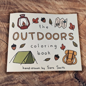 May include: A colouring book cover with a hand-drawn illustration of a tent, a backpack, a lantern, and acorns. The title is "The Outdoors Colouring Book" and the text at the bottom says "hand-drawn by Sara Smith".