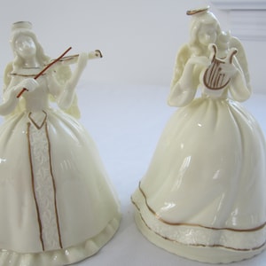 May include: Two white porcelain angel figurines, one playing a violin and the other a harp. Both angels are wearing gold trim and have a white background.